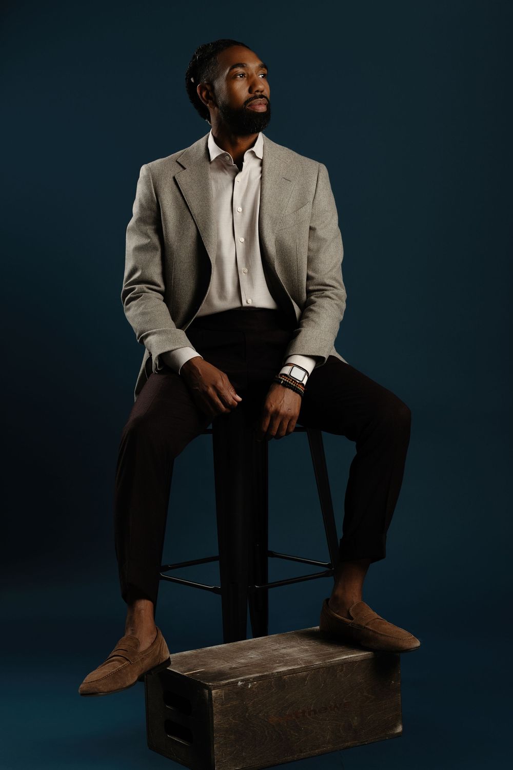 A stylish Black man in a gray blazer, white shirt, and black pants sits on a stool against a dark teal background.