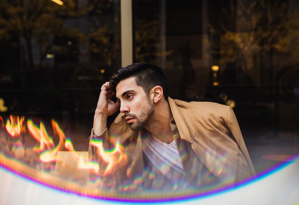 Young man in beige jacket resting head on hand, with dramatic fire reflections in foreground.