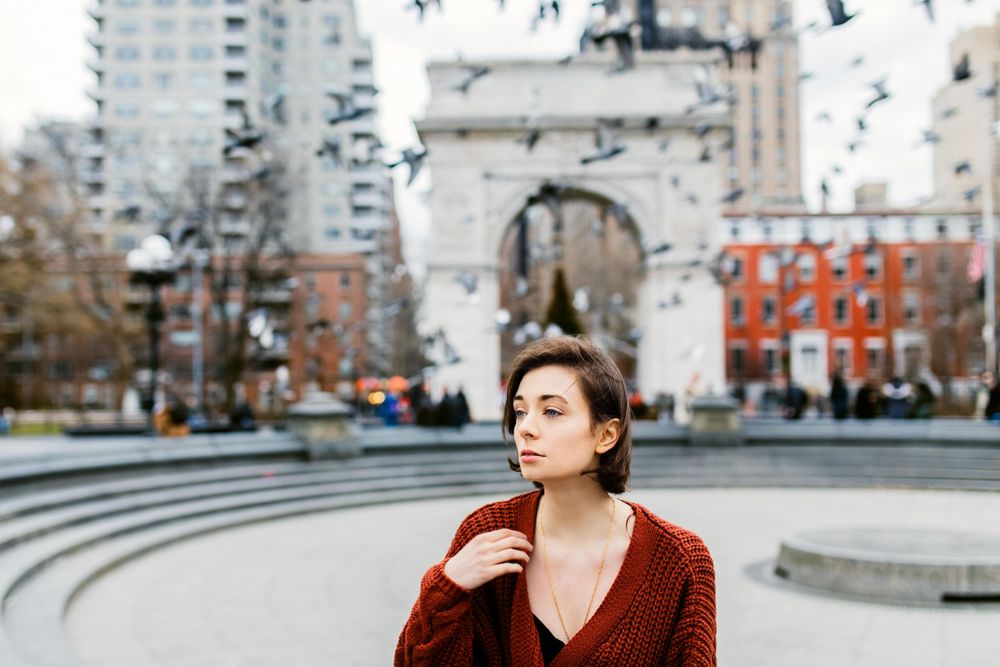 Young woman in red sweater stands near Washington Square Park arch with pigeons flying around her.