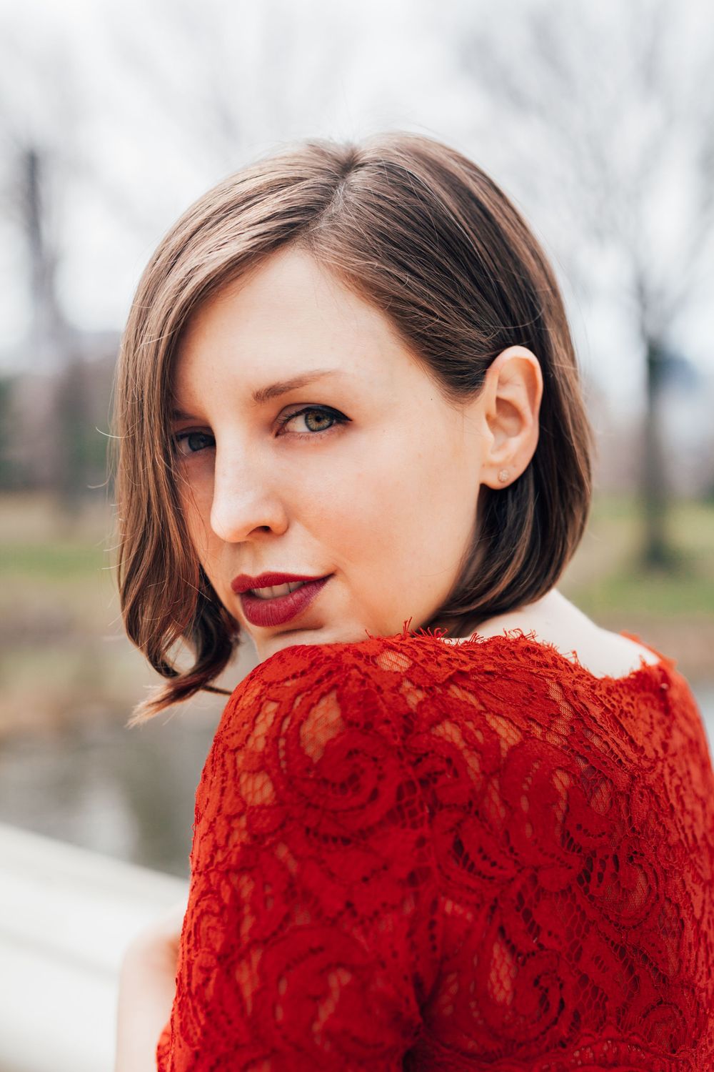 A young woman with a short bob hairstyle wearing a red lace dress poses outdoors with bare trees in the background.