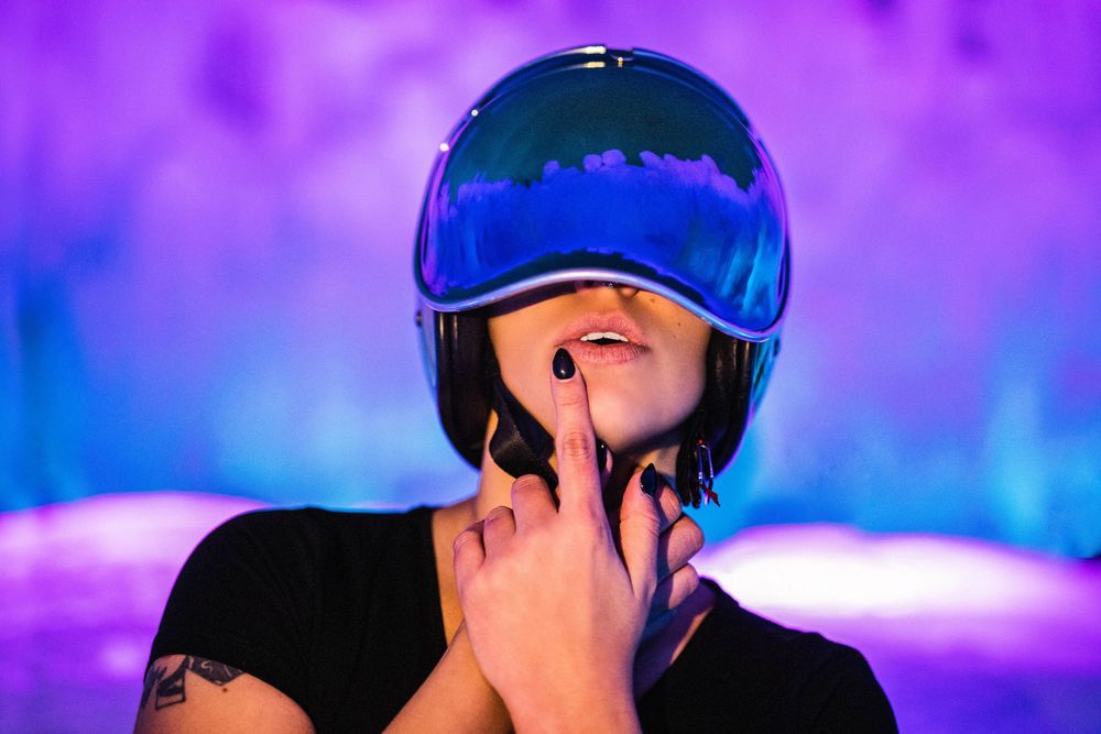 Person wearing a shiny blue helmet under purple neon lights, touching their chin with one finger.