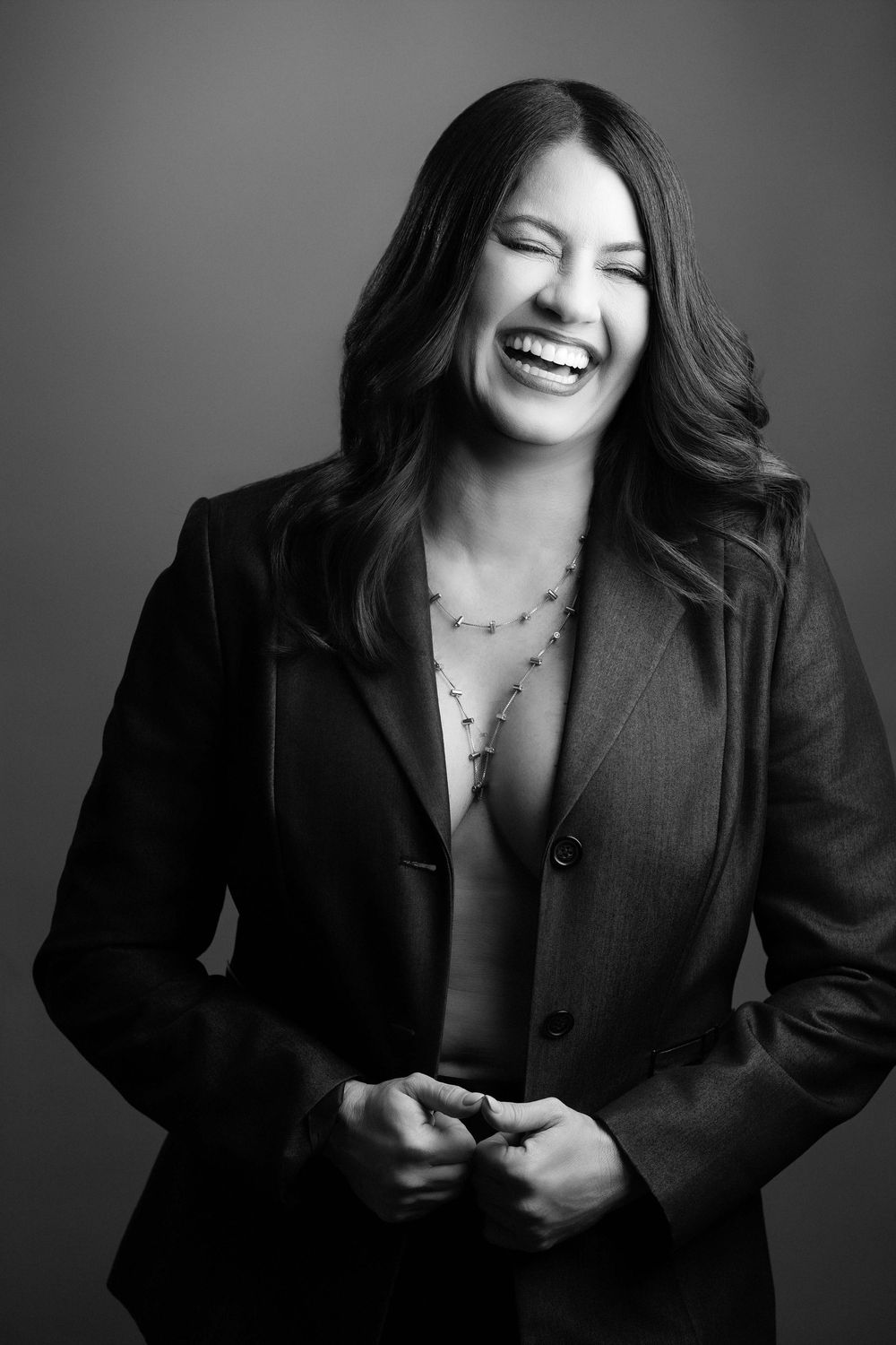 Black and white portrait of a laughing woman in a blazer and necklace, studio background.