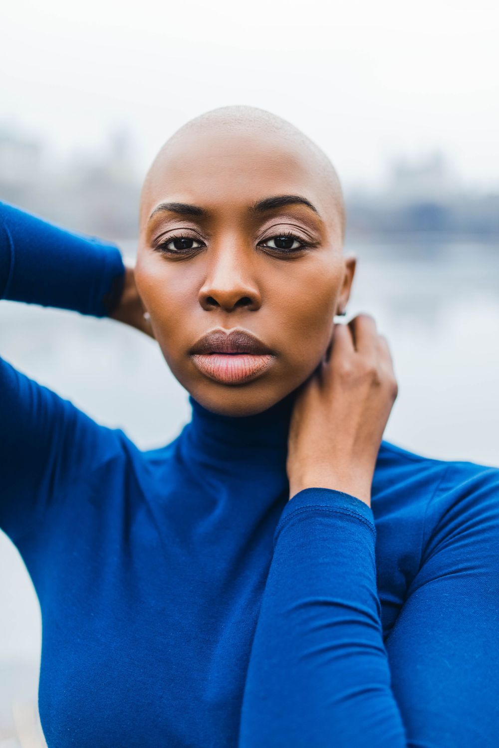 A person wearing a royal blue turtleneck poses with a bald head and striking makeup against a blurred outdoor background.