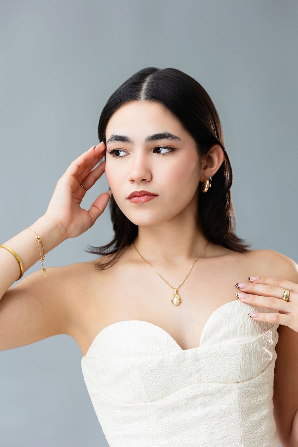 Young woman in white strapless top wearing gold jewelry including necklace, earrings, bracelet and ring on grey background.