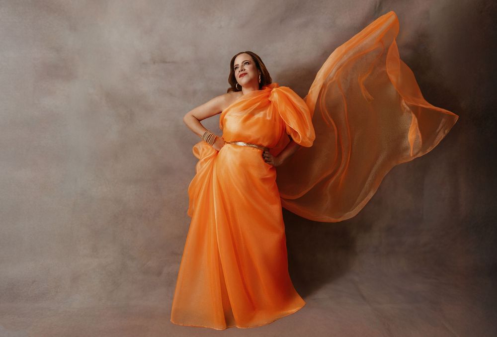 Woman in flowing orange chiffon gown with dramatic billowing fabric poses gracefully against neutral studio backdrop.