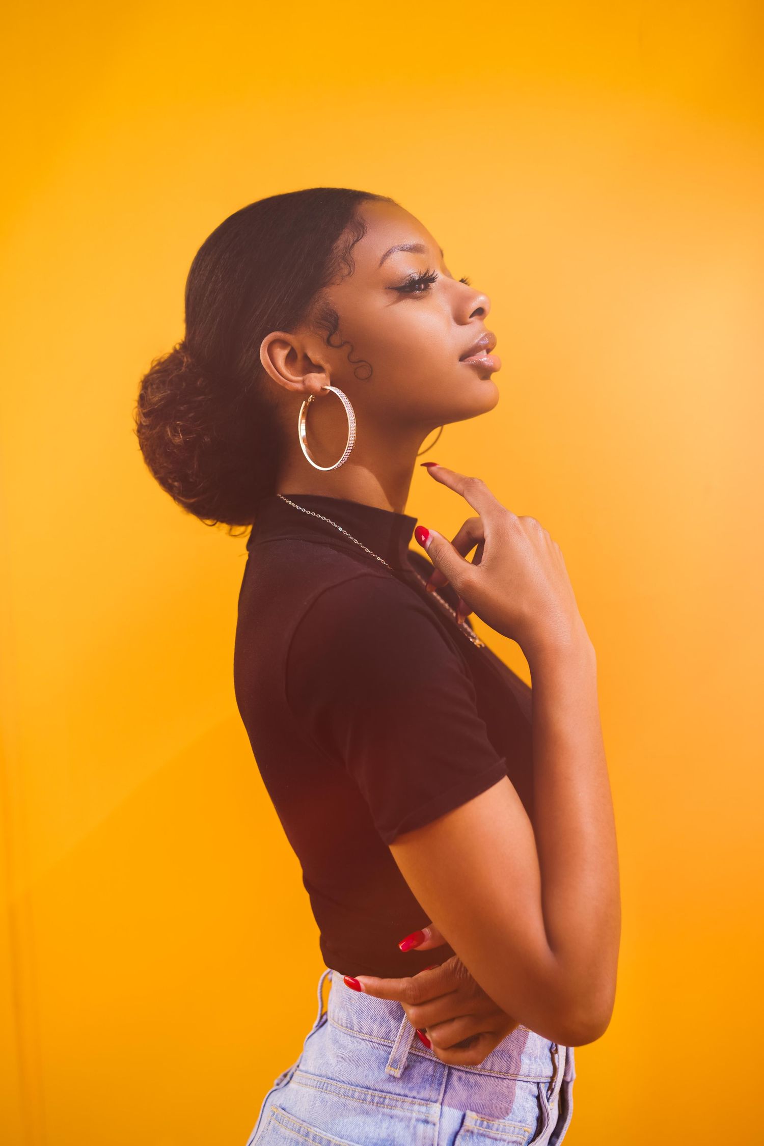 Stylish portrait against vibrant yellow background featuring silver hoop earrings, black crop top and light denim jeans.