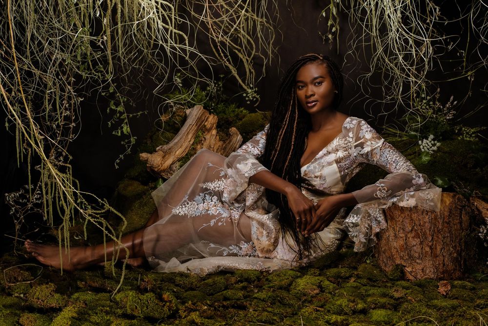 A Black woman in a floral dress reclines gracefully among lush green moss and hanging vines in a forest setting.