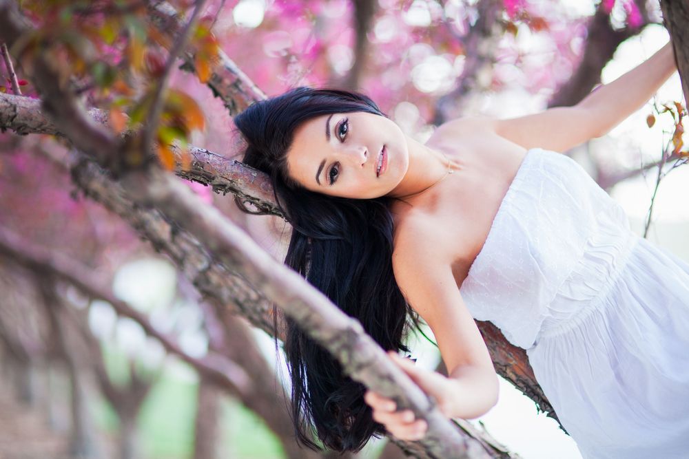 Young woman in white dress leaning on cherry blossom tree branches, smiling softly among pink spring flowers.