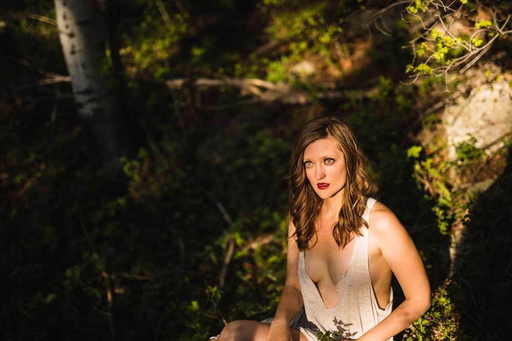 Woman in white dress sitting outdoors among lush green trees and dappled sunlight in a forest setting.