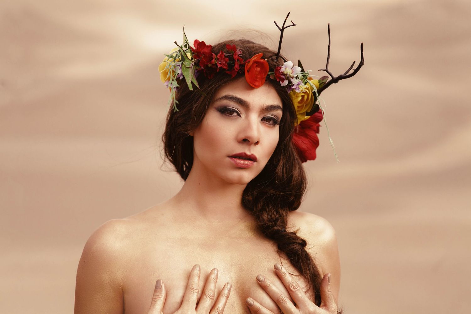 Artistic portrait showing floral crown with red roses and antlers against warm beige background.