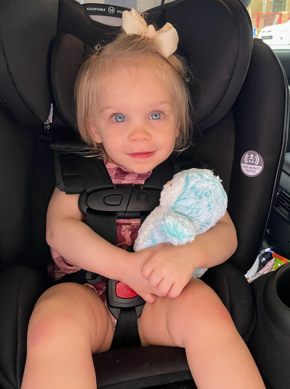 A young child secured in a black car seat holding a light blue plush toy while smiling.