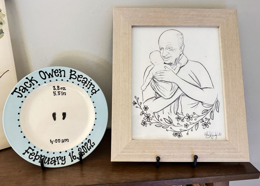 A framed pencil sketch artwork displayed next to a decorative white ceramic plate with blue text detailing.