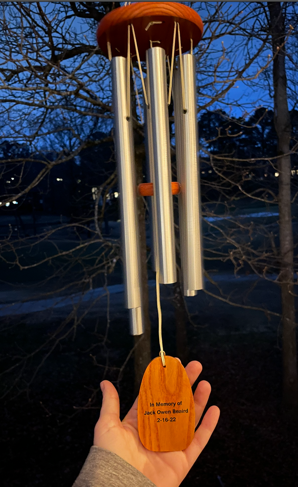 A wooden wind chime with silver metal tubes hangs against a blue sky and bare tree branches at dusk.