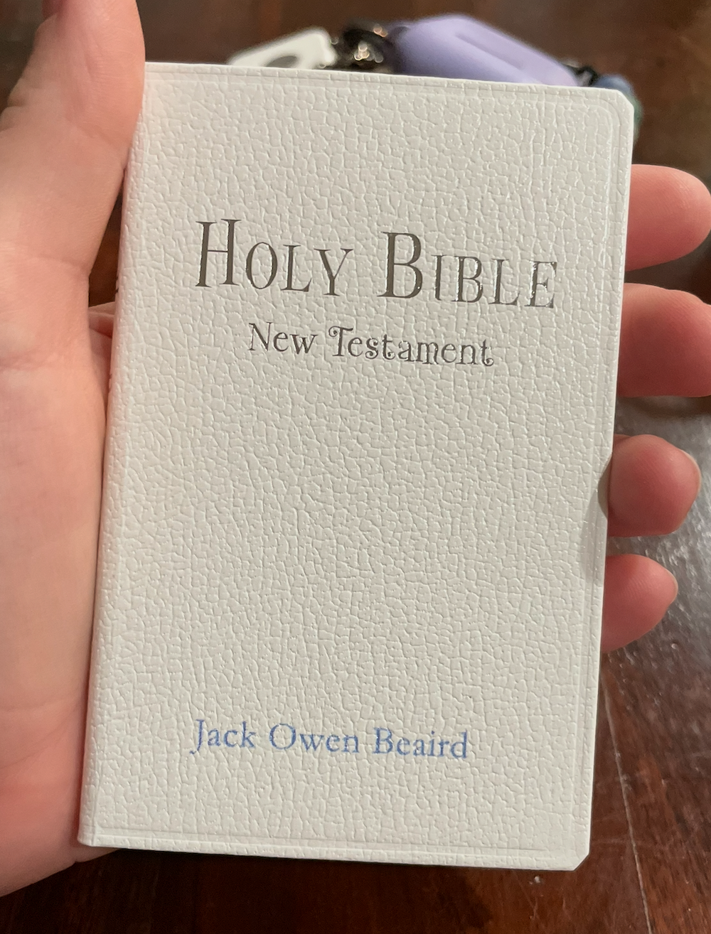 A white Holy Bible New Testament book with embossed text and a name on the cover is held in someone's hand.