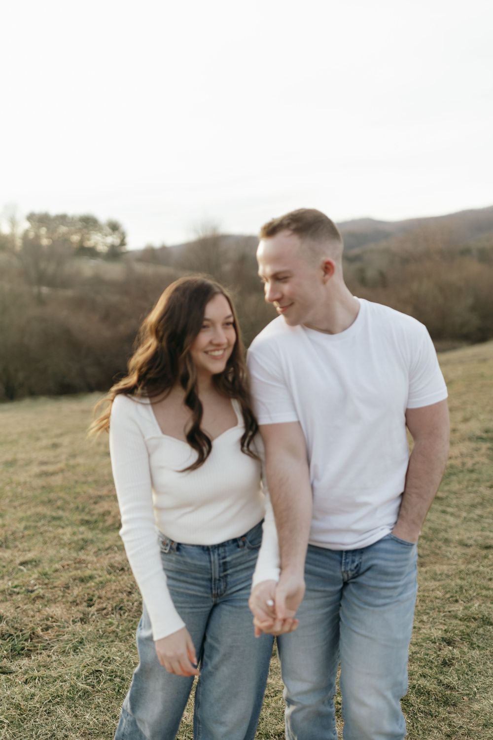 Couple in coordinating white tops and jeans share loving glance in grassy field.