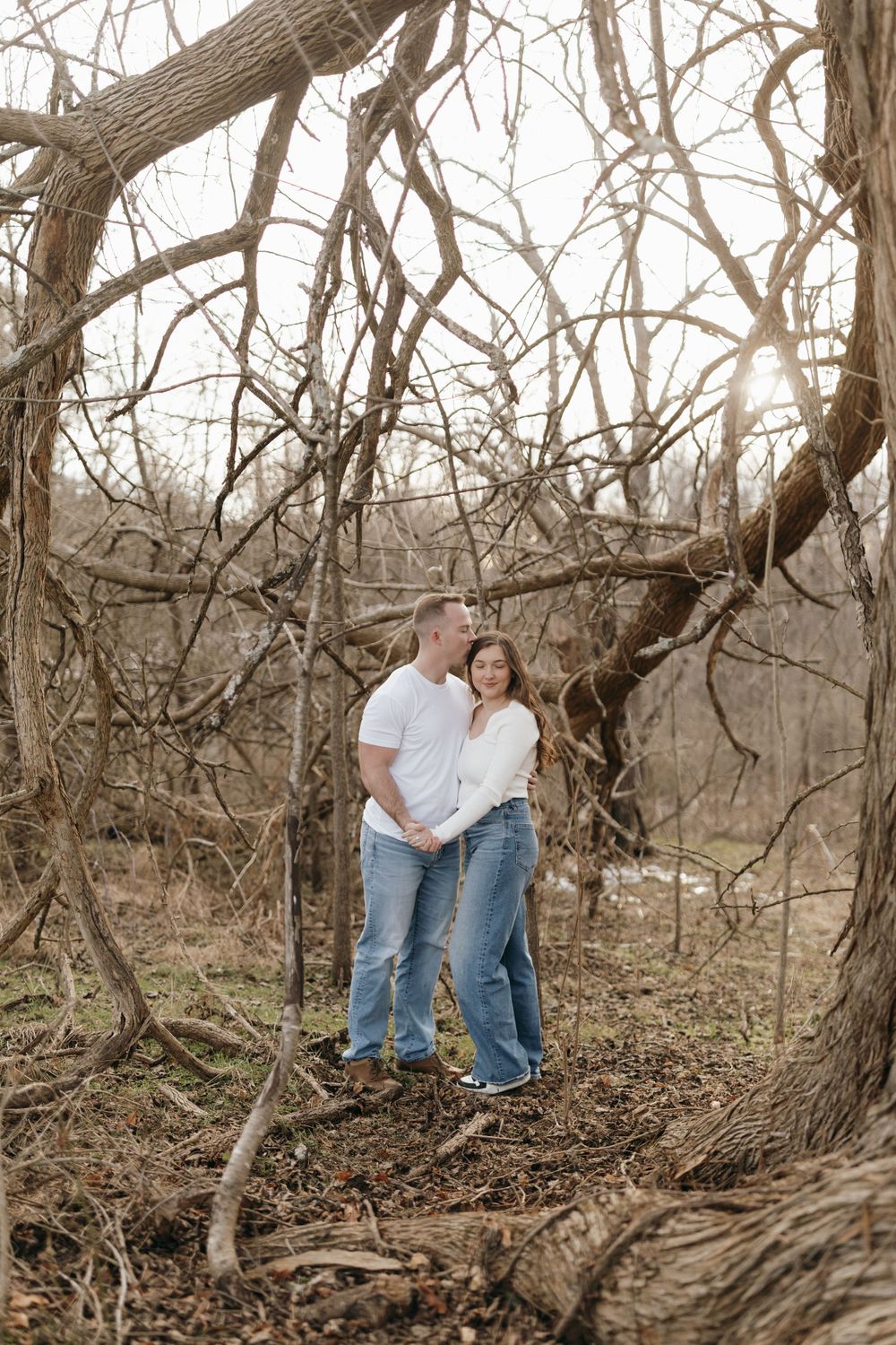 A couple stands together among bare winter trees in a wooded setting.