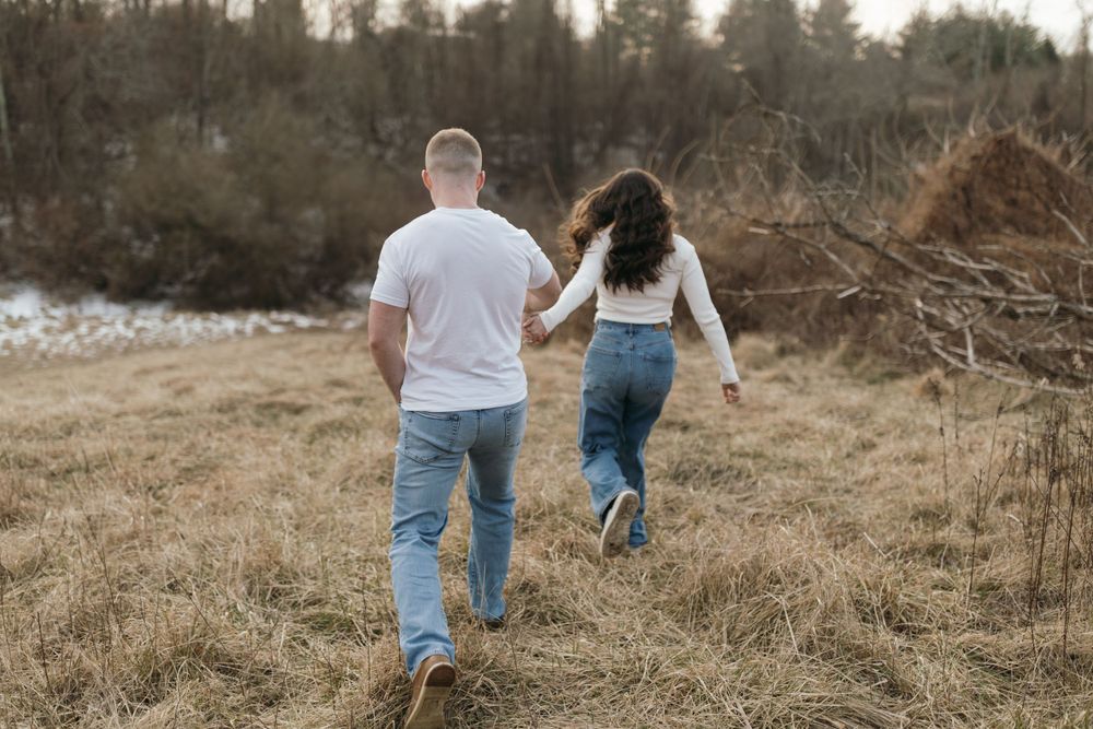 A couple runs playfully through a field during a sunset photoshoot.