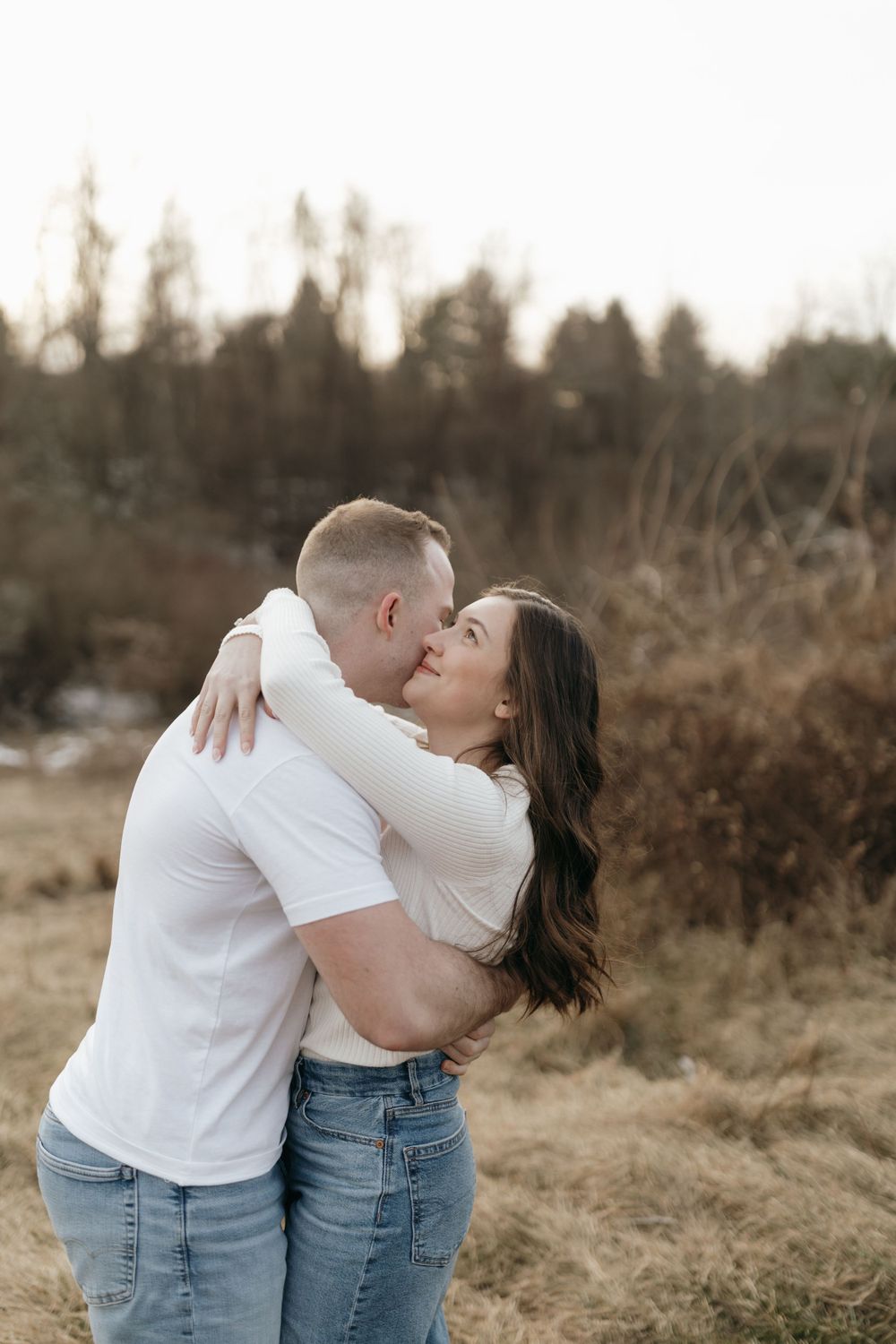 A couple shares an intimate embrace in a winter field with trees in the background.