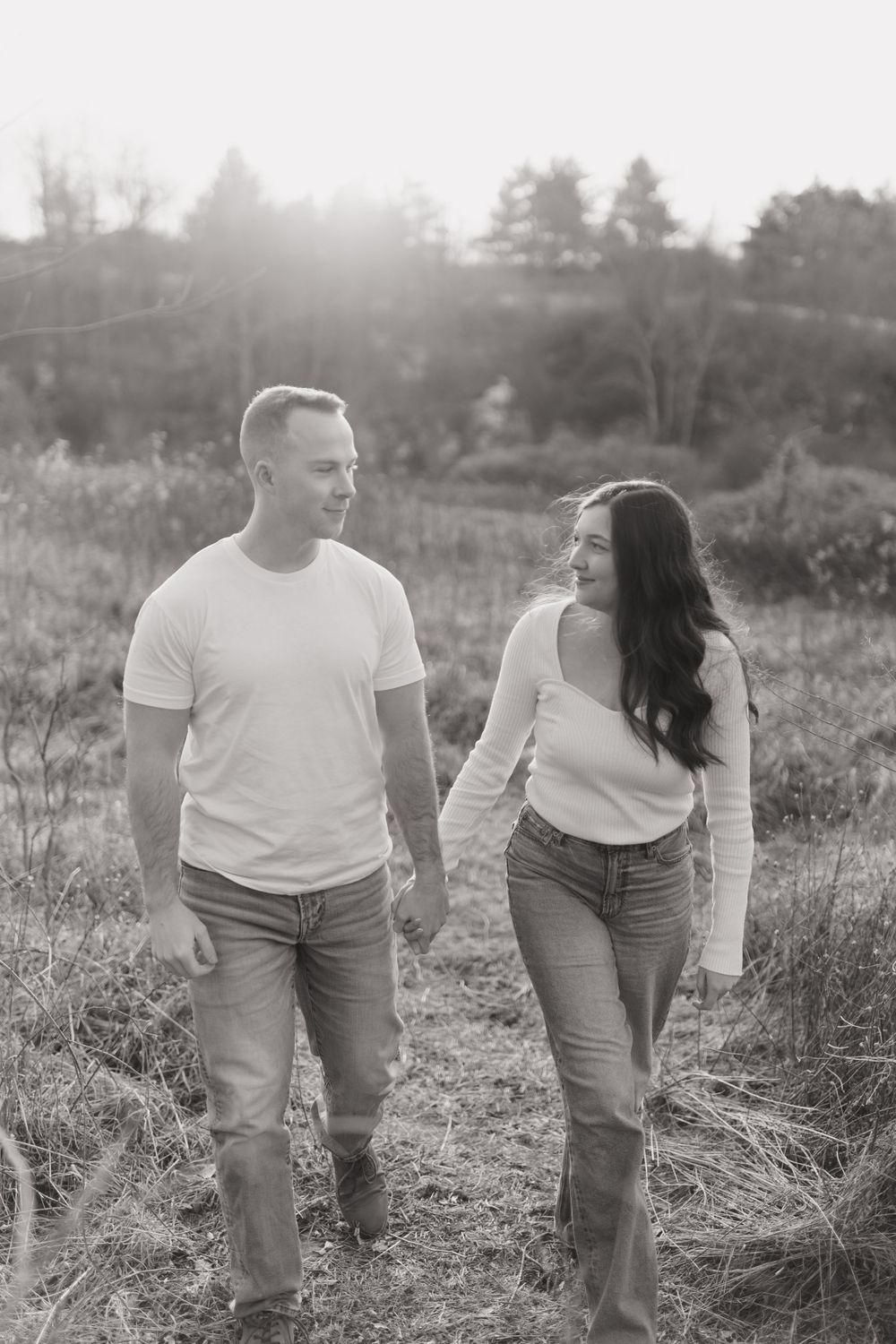 Black and white photo of couple walking together in field with sunlit backdrop.