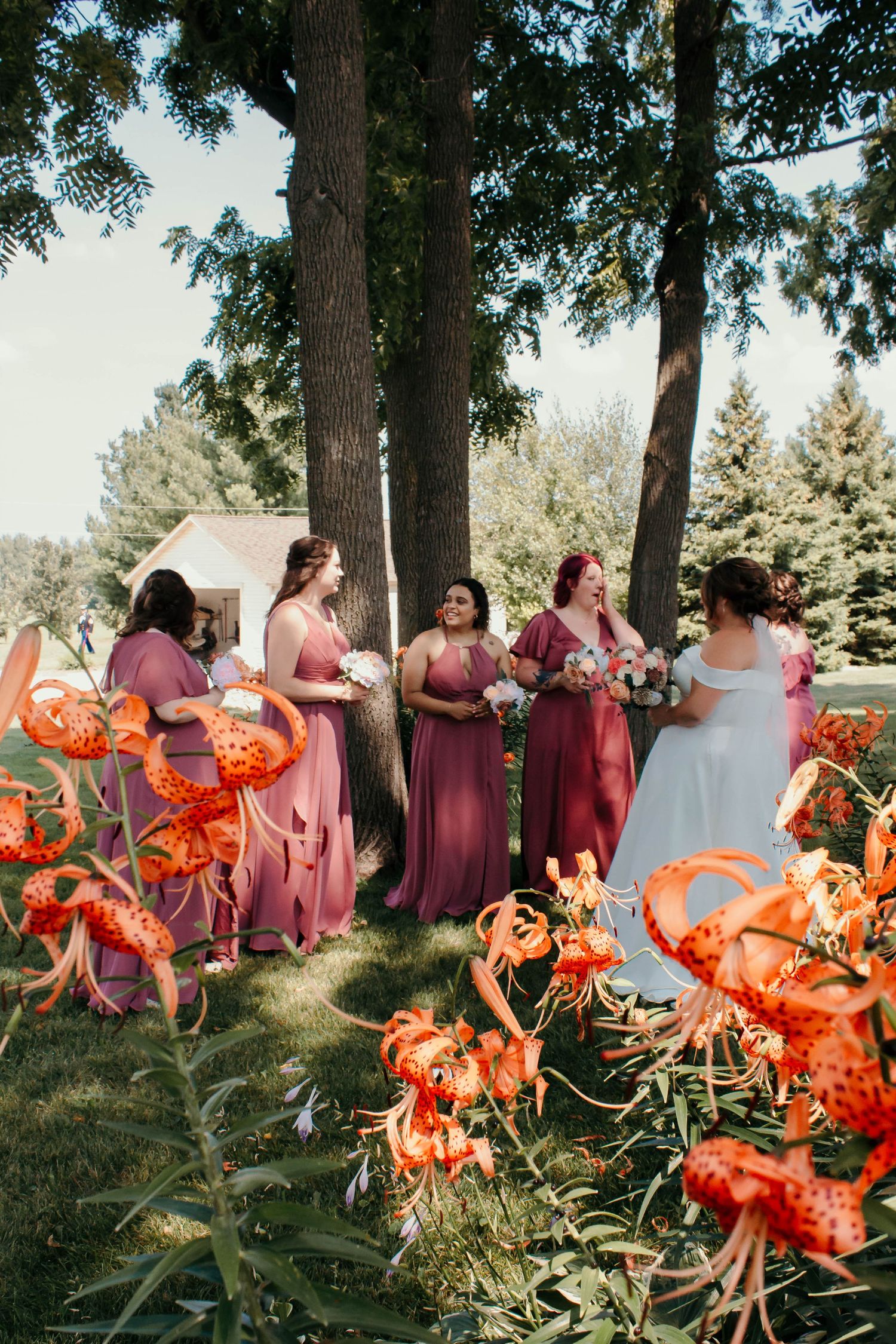 Wedding party in pink and coral dresses poses outdoors with vibrant tiger lilies in the foreground.