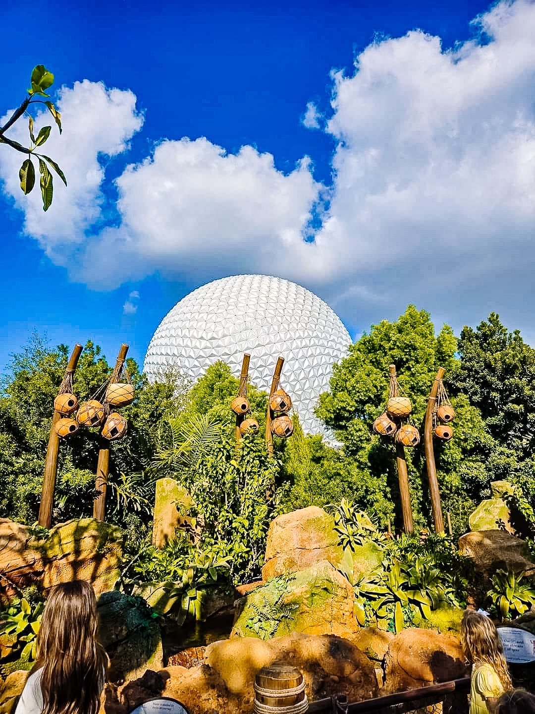 Rock formations and landscaping surround the iconic Spaceship Earth geodesic sphere at EPCOT under blue skies.