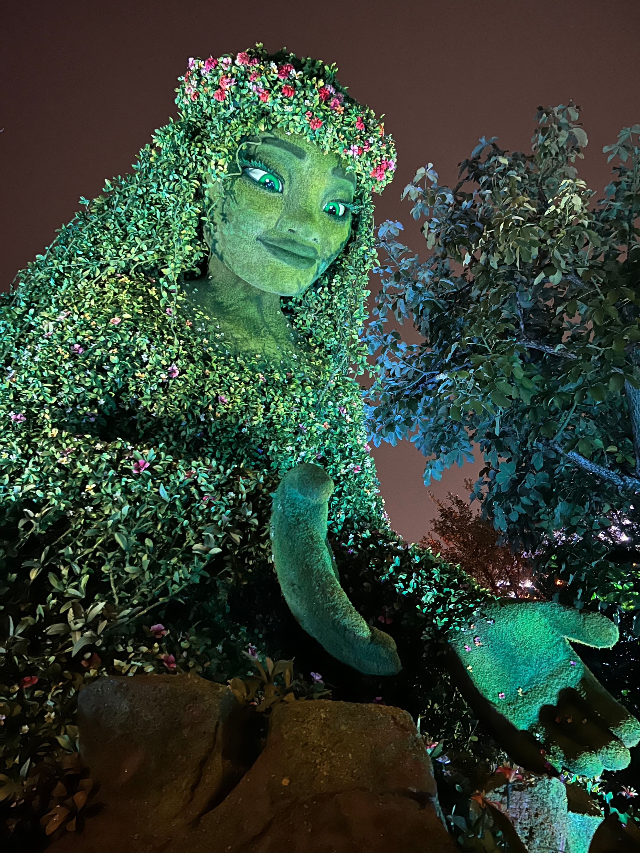 A large green garden sculpture lit up at night shows a nature-inspired figure emerging from foliage and branches.