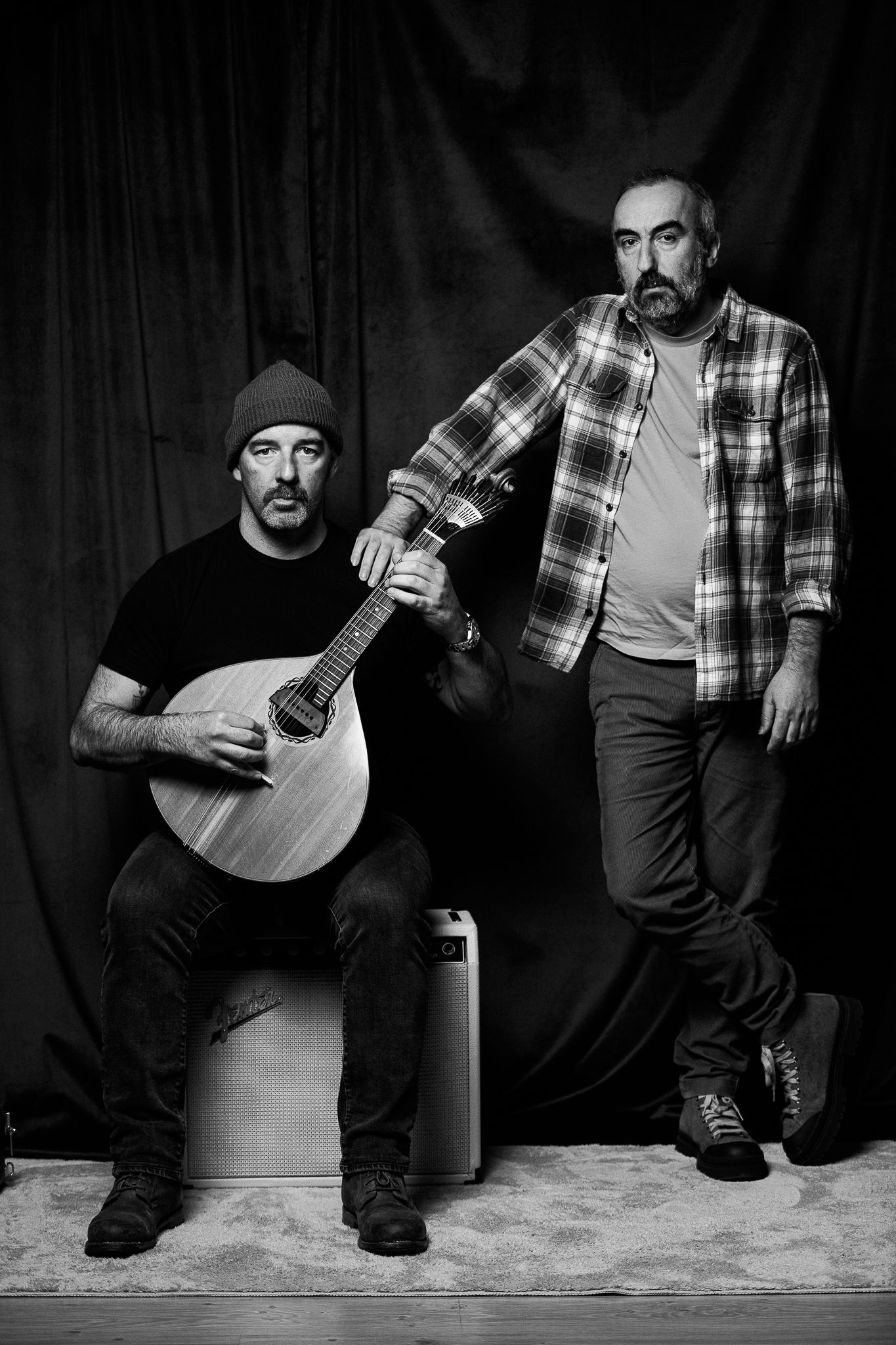Two musicians pose in black and white photo - one seated with banjo, one standing, both wearing casual attire.