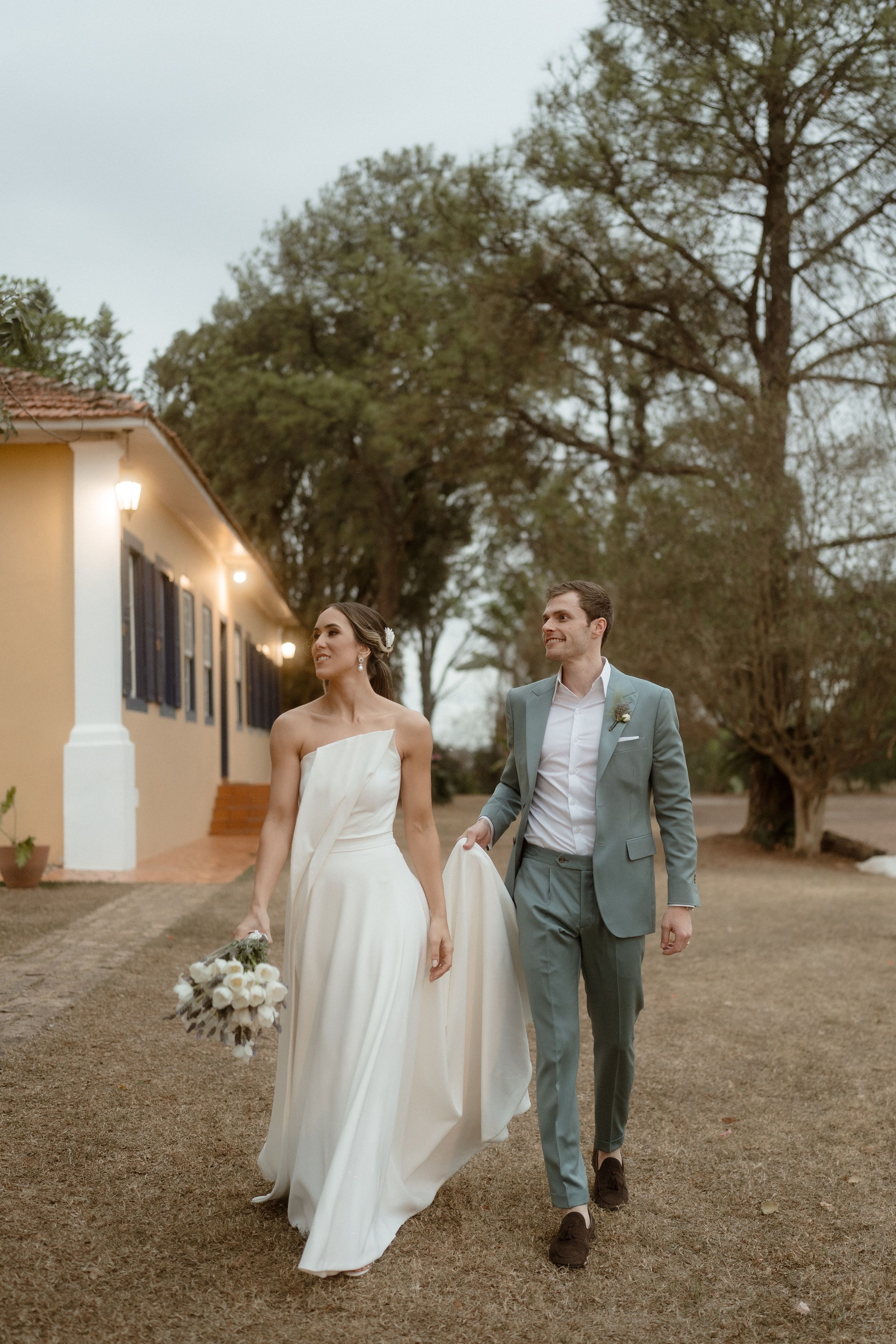 A romantic couple walks together on a garden path at dusk in front of a yellow house with string lights.