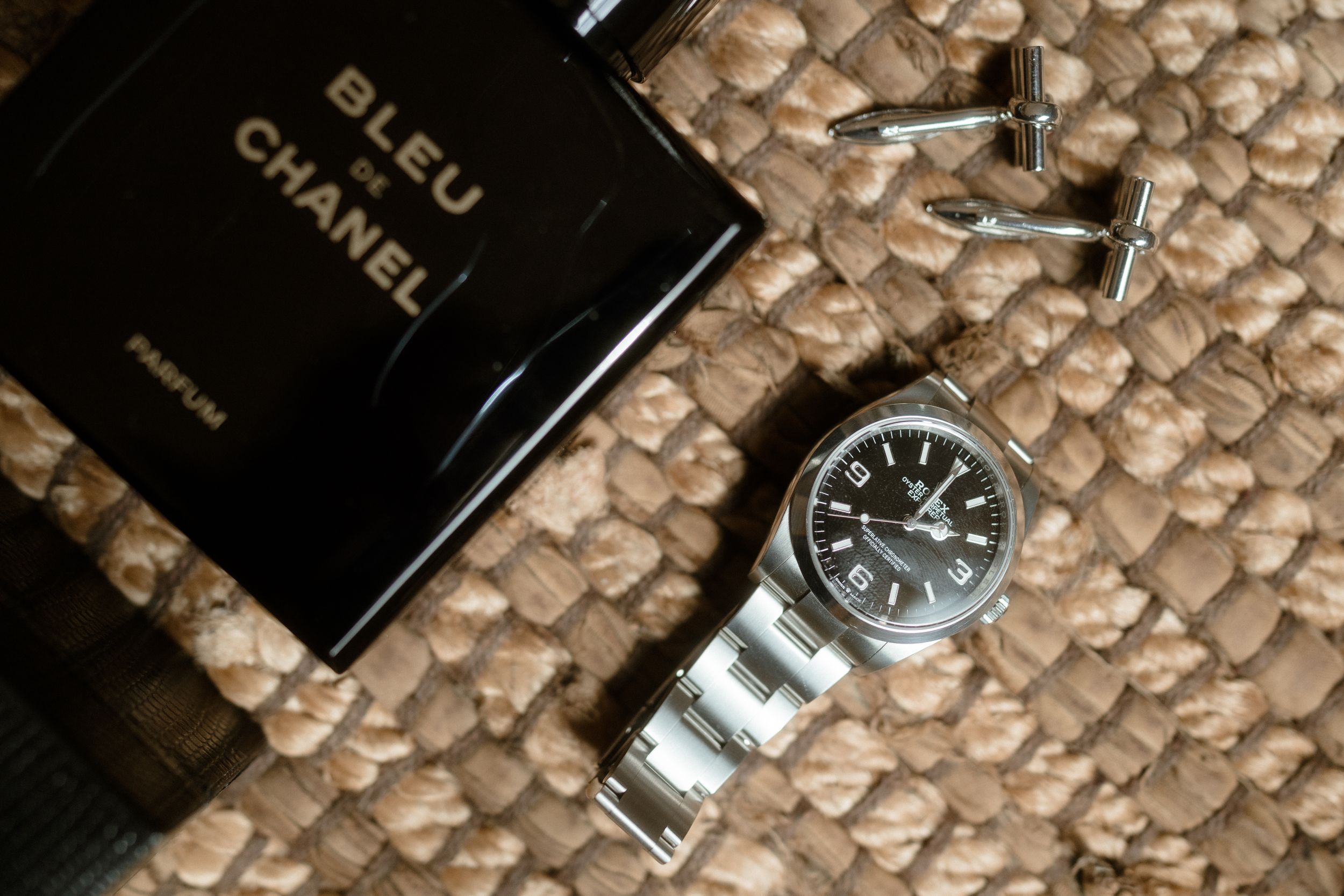 Luxury Chanel perfume bottle and silver watch displayed on textured beige fabric.