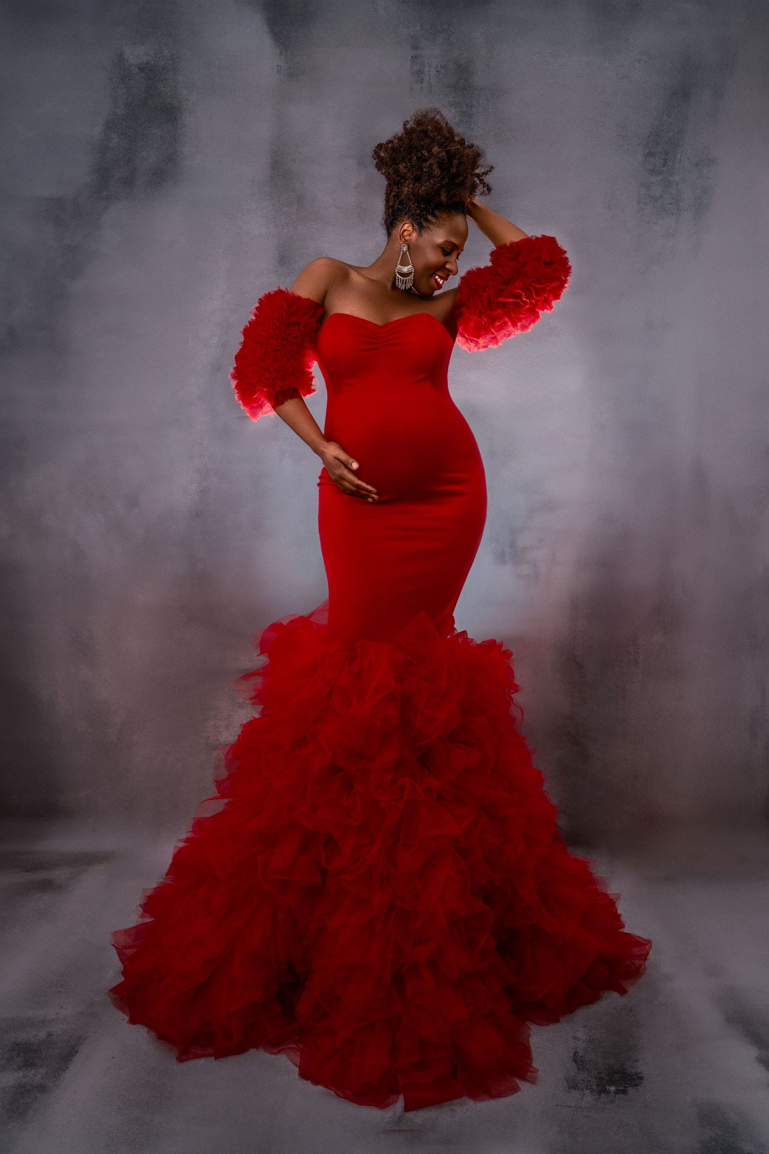 A maternity photo shoot wiith Dramatic red mermaid gown with ruffles photographed against a moody grey background.