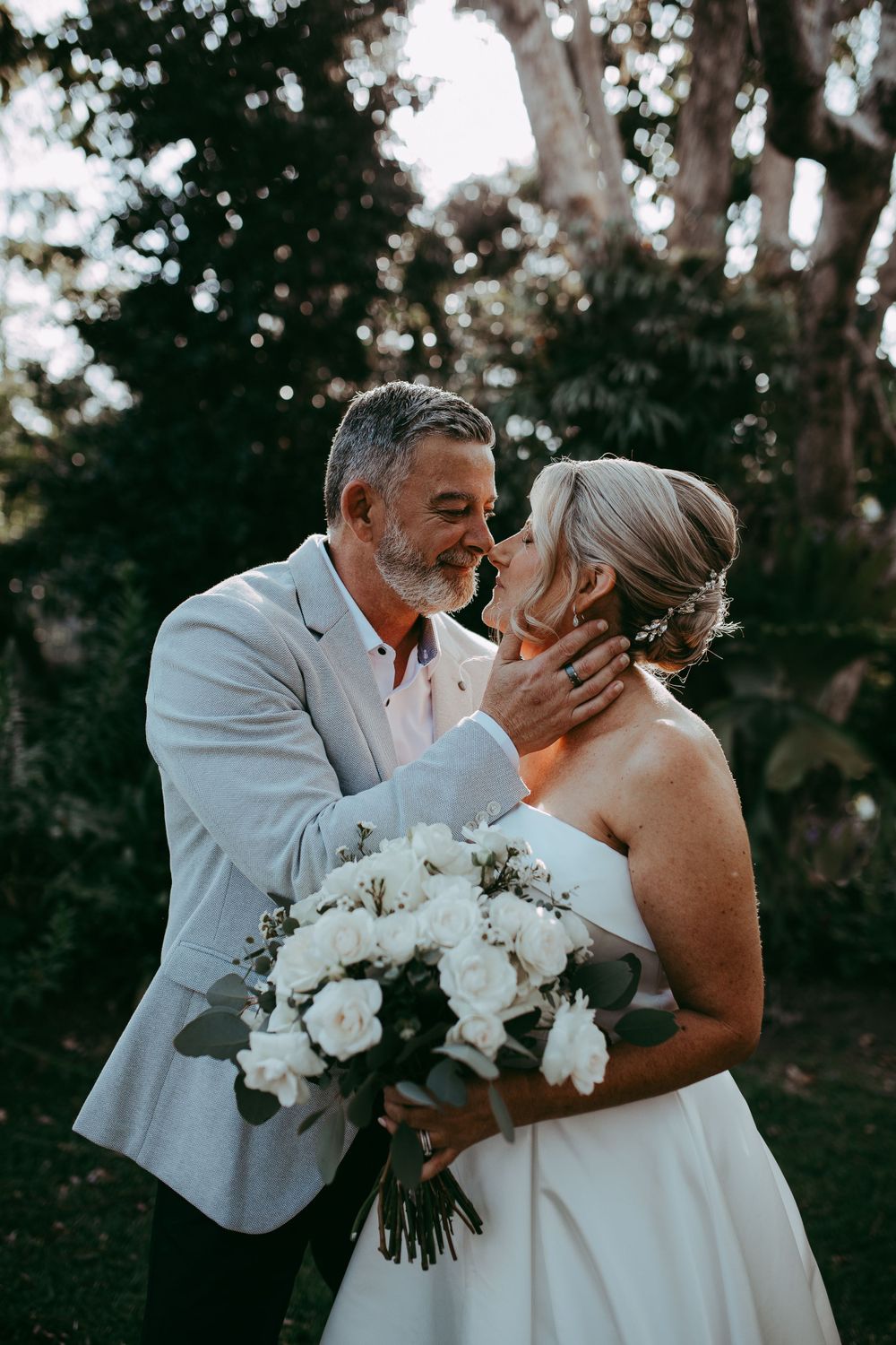 Romantic outdoor wedding moment with couple embracing and holding elegant white rose bouquet at sunset.