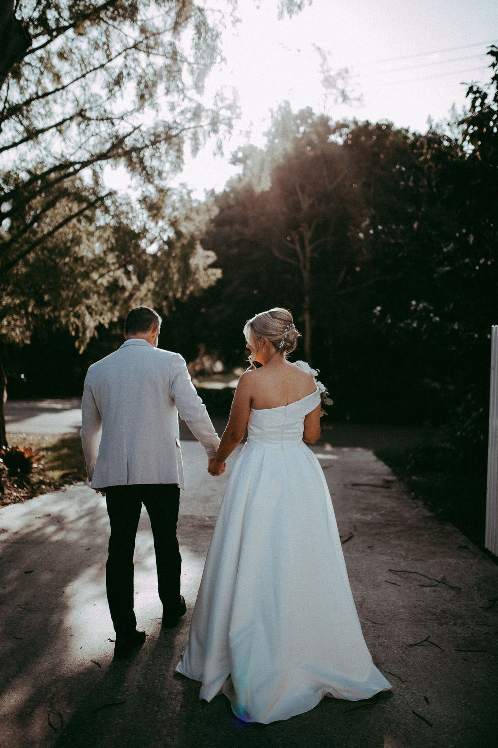 A couple holding hands while walking down a sunlit path, the bride wearing an elegant one-shoulder white gown.