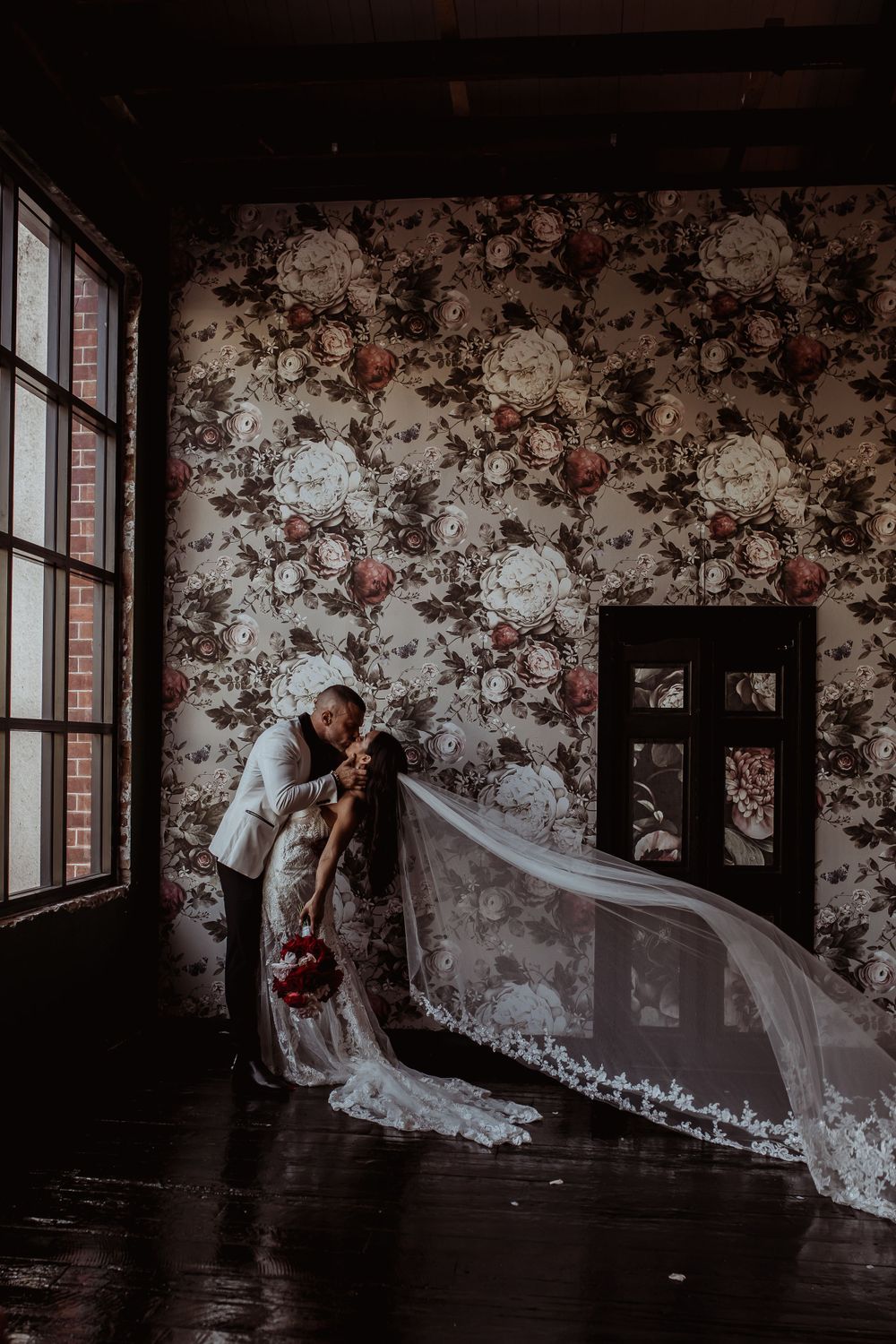Romantic wedding moment with fluttering veil against vintage floral wallpaper in moody indoor setting by large windows.