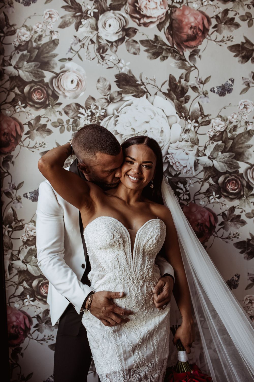 Elegant wedding portrait against a floral wallpaper backdrop featuring a white lace dress and flowing veil.