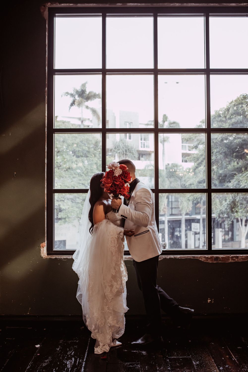 A couple in wedding attire embraces romantically in front of a large industrial window with red flowers.