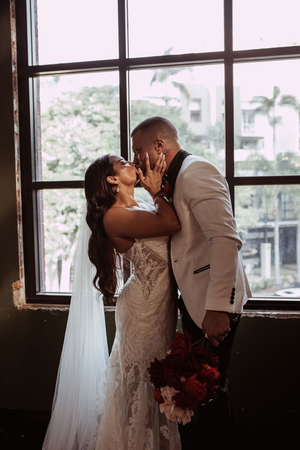 A romantic wedding portrait shows the couple sharing a kiss by a large window with palm trees visible outside.