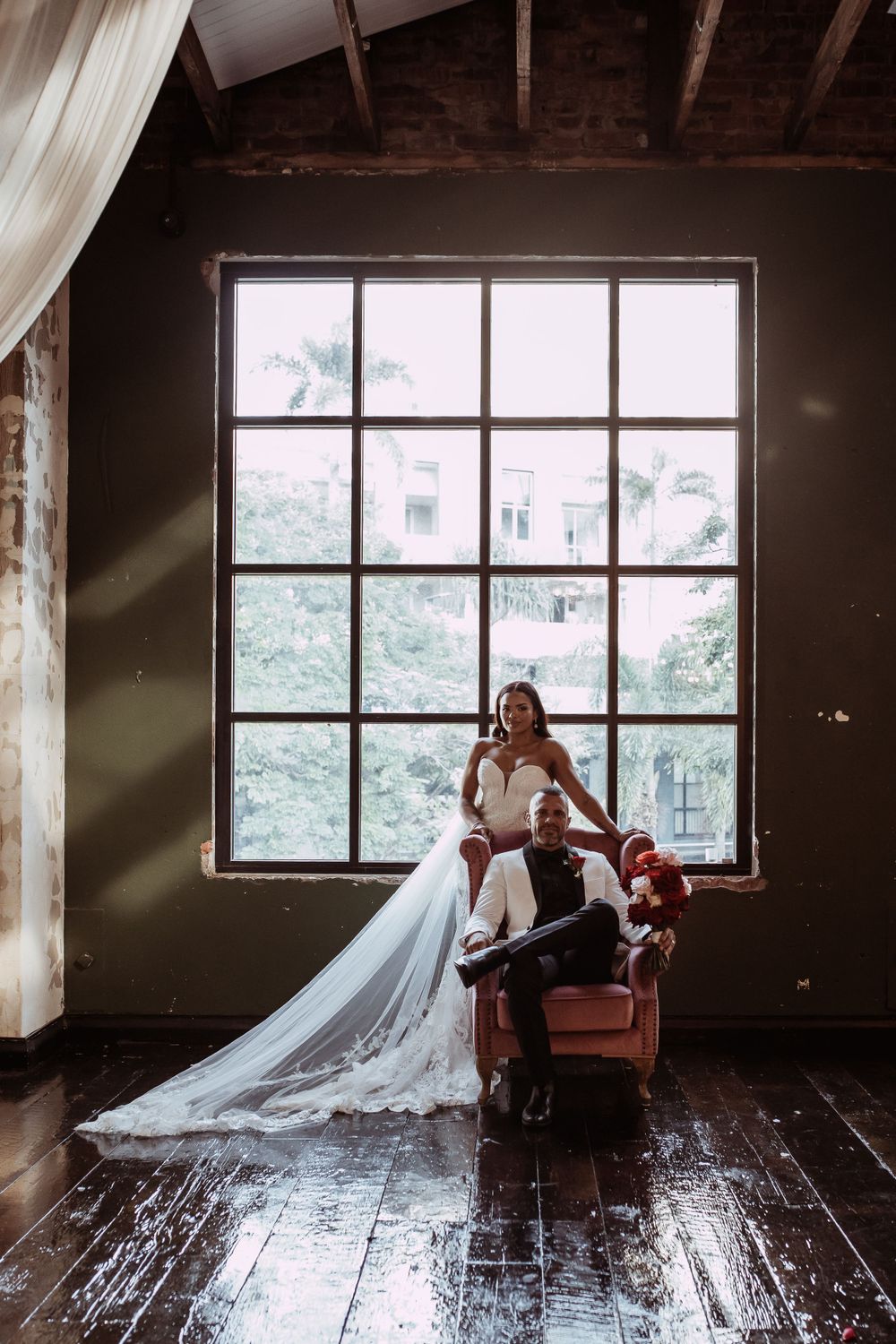 Moody wedding photo in rustic room with large window, vintage armchair and flowing white dress on wooden floor.