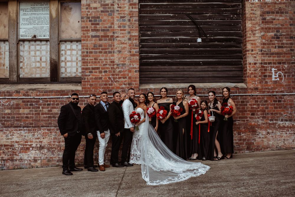 Wedding party poses together against rustic brick wall with bride's long veil trailing behind.