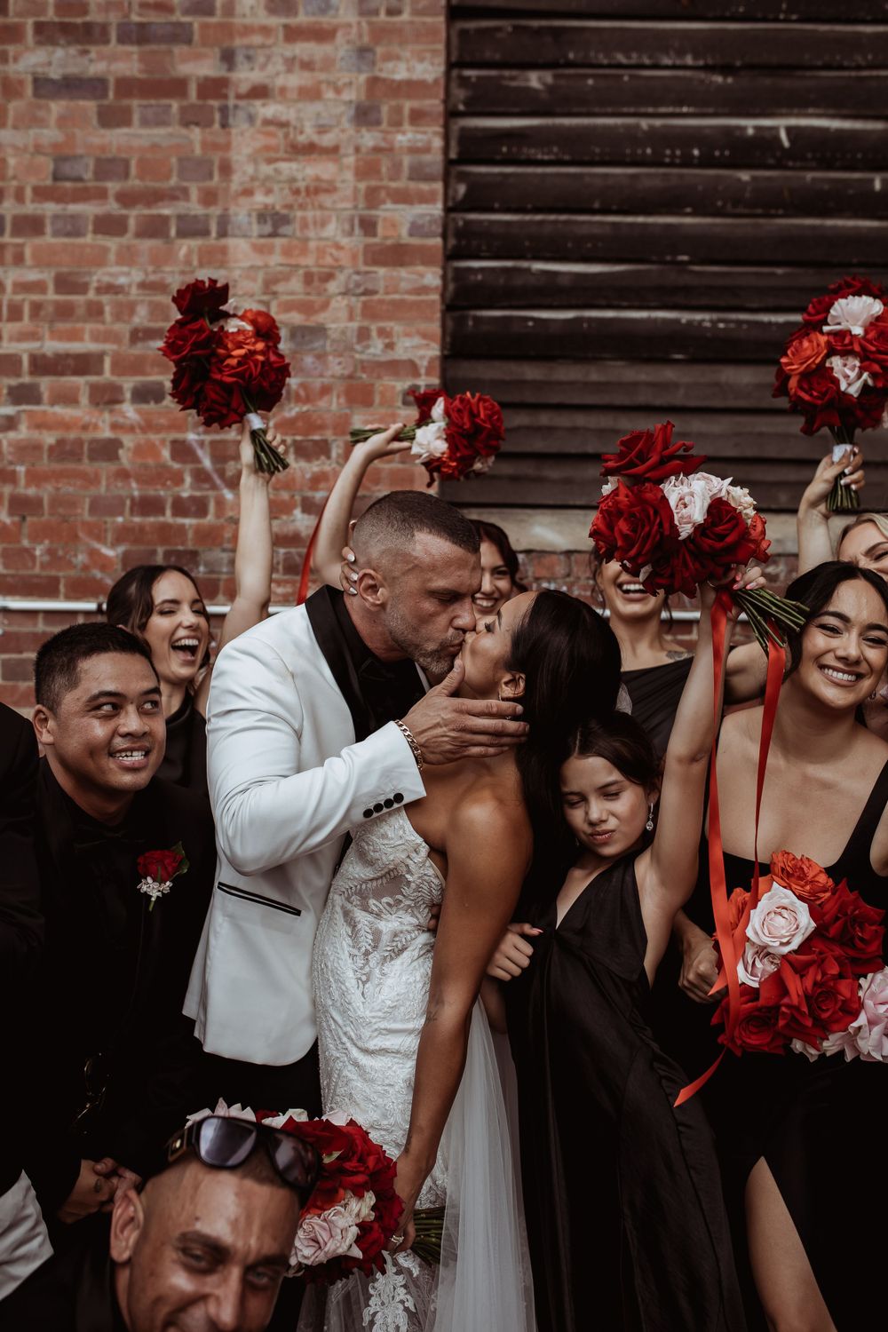 A wedding celebration with bridesmaids and groomsmen holding vibrant red bouquets against exposed brick wall.
