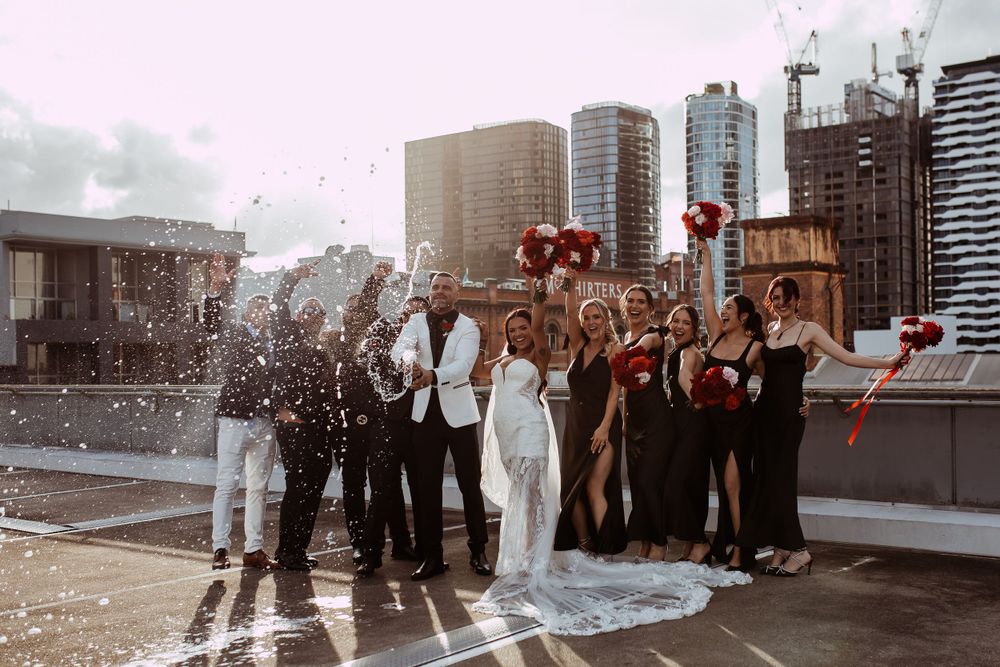 A celebratory group pops champagne on an urban rooftop with city skyline views at sunset.