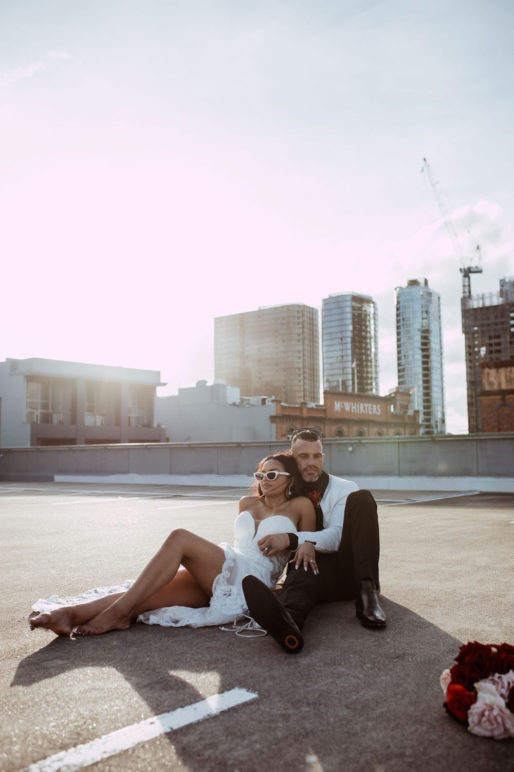 A romantic couple relaxes together on an urban rooftop with city buildings and sunset in the background.