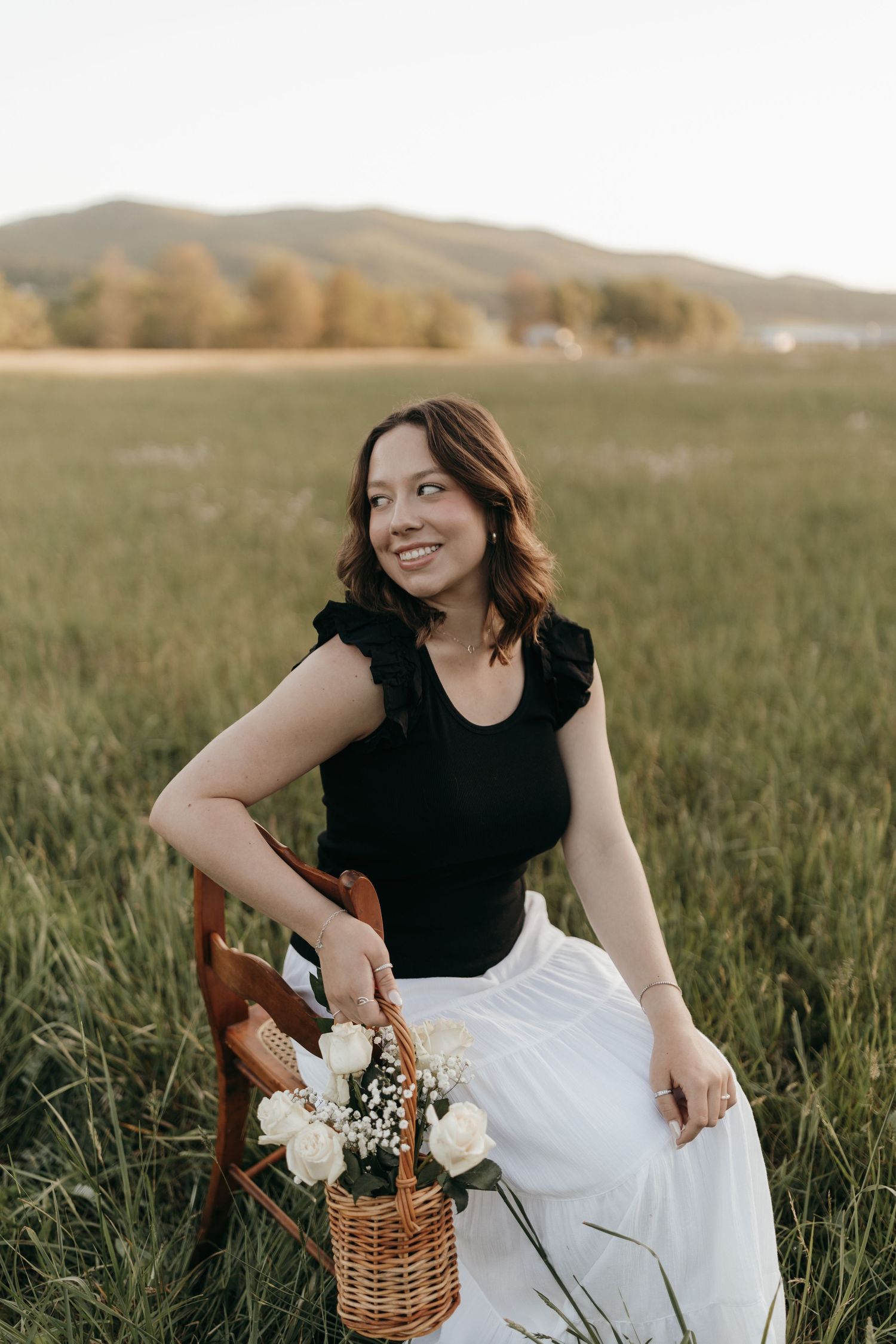 A woman in a white skirt sits with a wicker basket of flowers in a mountain meadow at golden hour.  Virginia graduation/senior session.