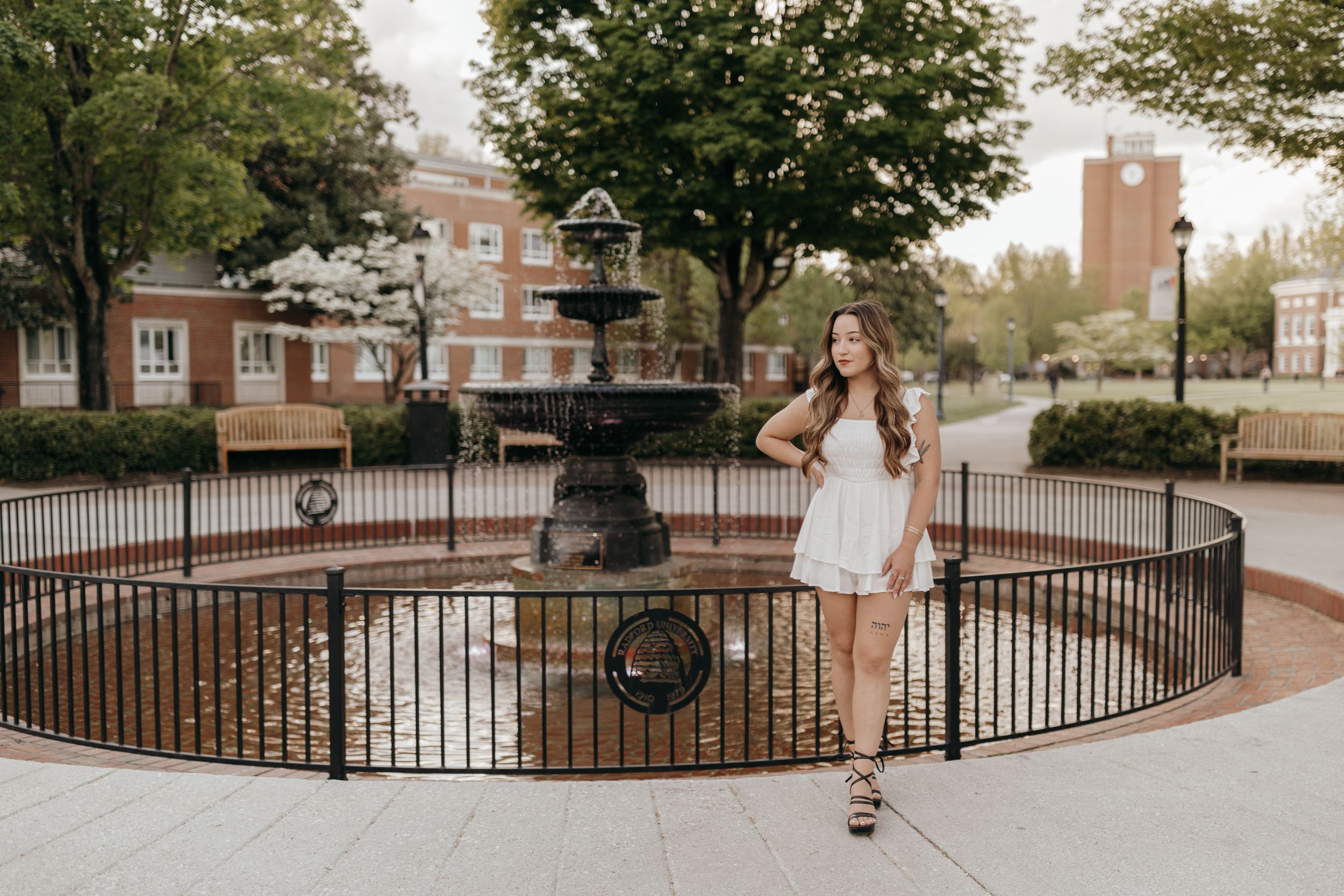 Graduate photographed next to ornate iron fence and fountain in white dress on university grounds. Virginia graduation/senior session.