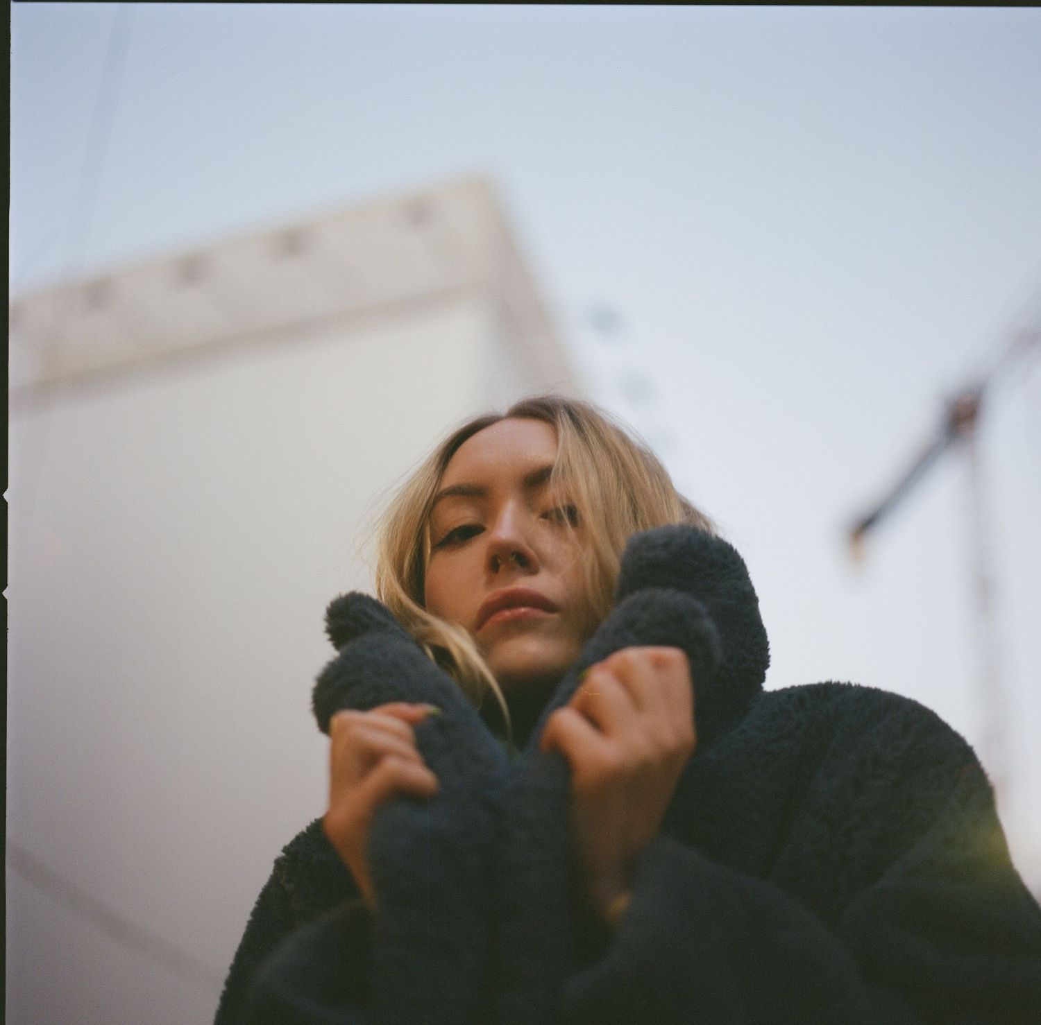 Person in black winter coat with blonde hair against cloudy sky in moody portrait.