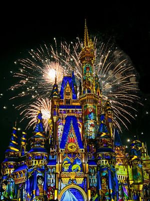 Fireworks burst behind Cinderella Castle illuminated in blue and gold lights during Disney's nighttime spectacular show.