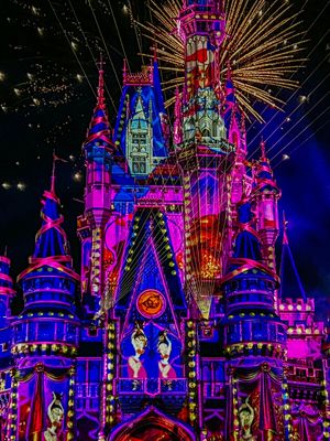 Cinderella Castle at Disney World illuminated in vibrant pink and blue lights with fireworks bursting behind it at night.