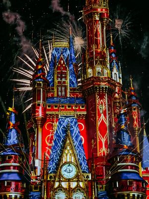 Illuminated Disney castle at night with red and blue lights and fireworks bursting in the dark starry sky.