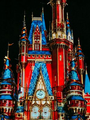 Illuminated Cinderella Castle at Disney World glows in vibrant red and blue colors with ornate spires and clock tower at night.