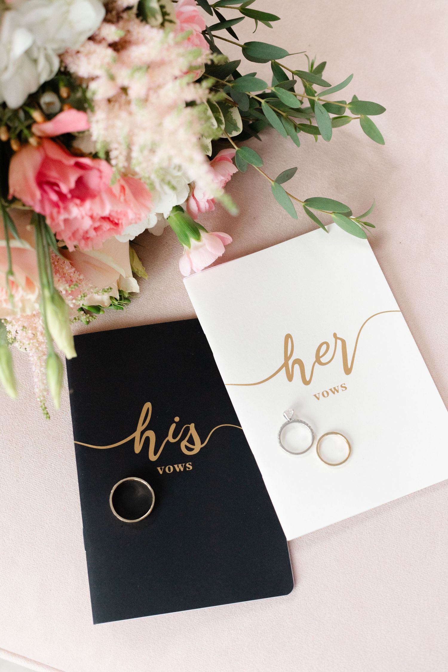 Wedding vow books in black and white with gold lettering display his and her vows alongside wedding rings and pink flowers.