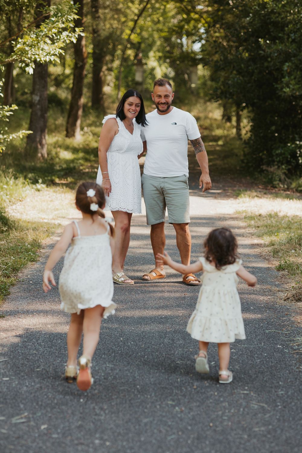 Family of four enjoying a sunny walk in a tree-lined park, two young girls running toward smiling parents.